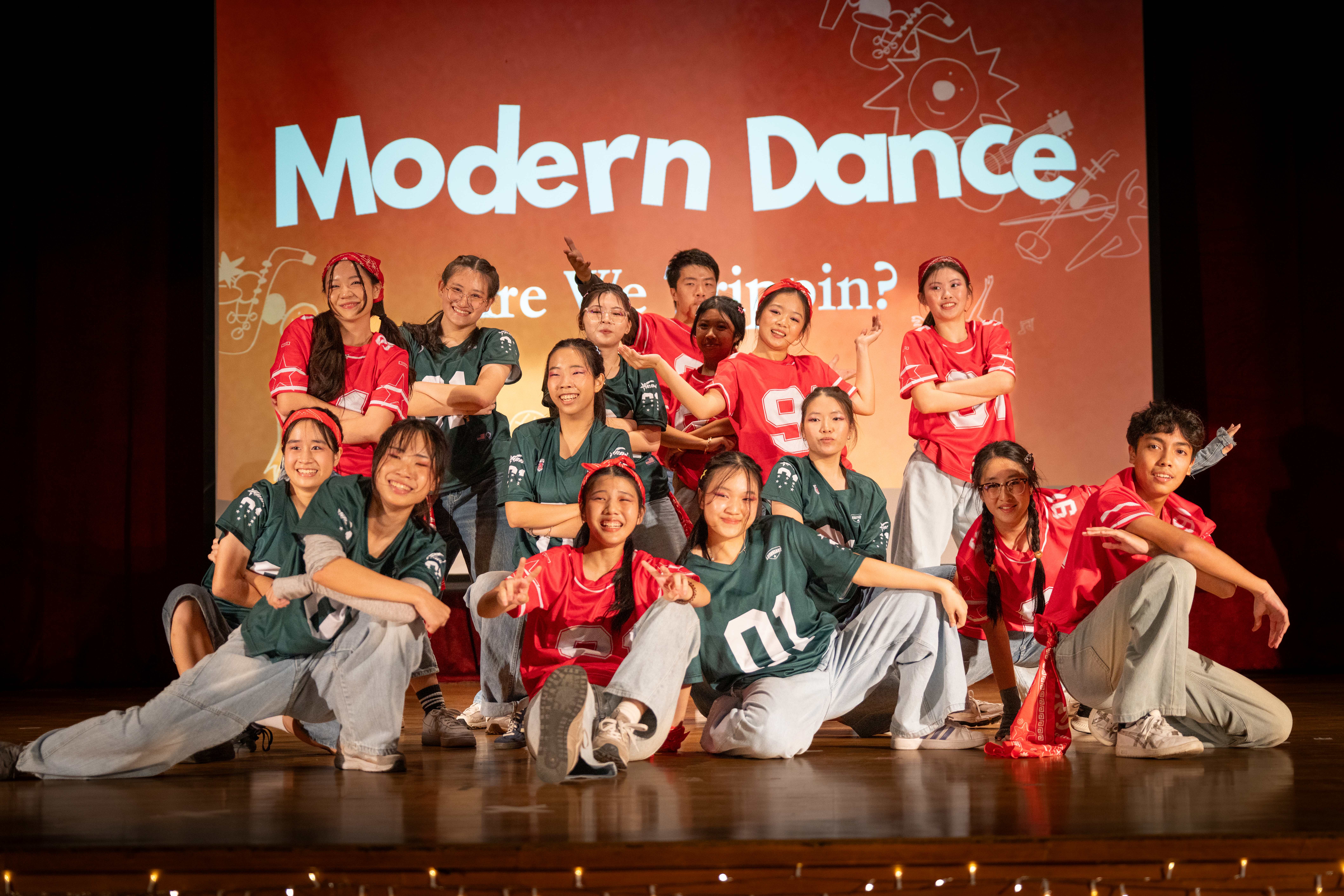A cheerful dance group in red and green jerseys poses on stage after performing "Are We Tripping?", smiling at the camera.
